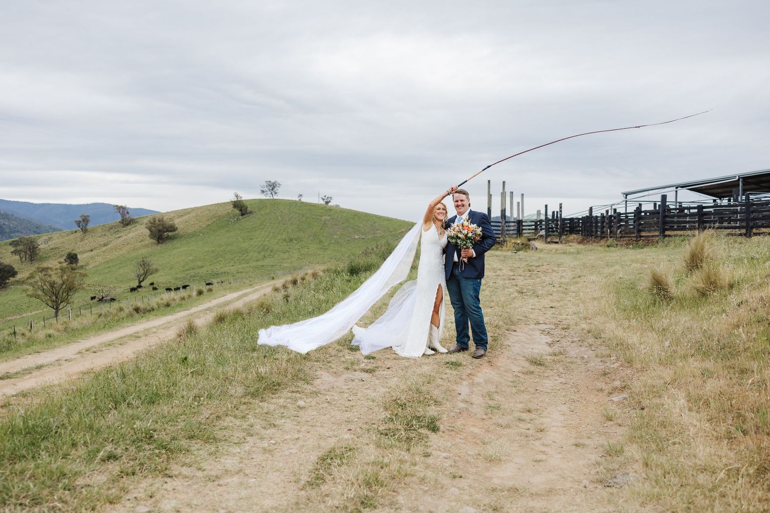 Two people embrace on a scenic hilltop path with rolling green hills and cloudy skies in the background.
