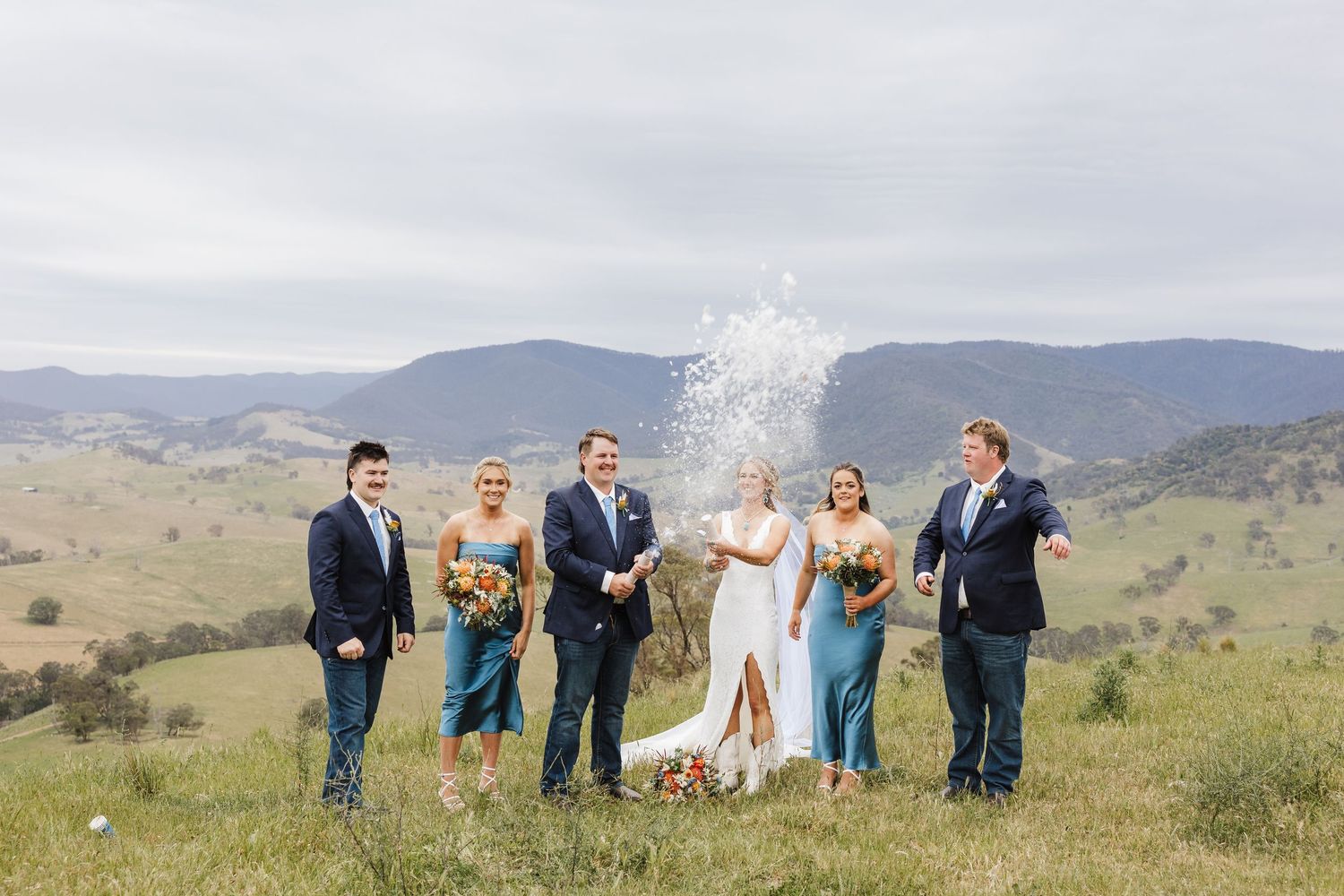 Wedding party celebrates with champagne spray against backdrop of rolling hills and mountains on a cloudy day.