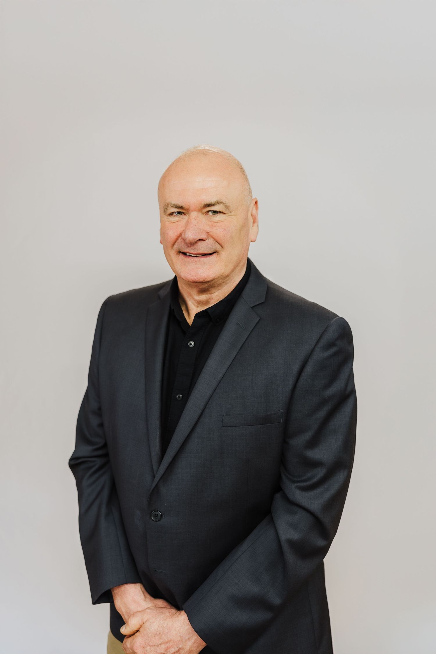 Professional headshot of someone wearing a black suit and shirt against a light gray background.