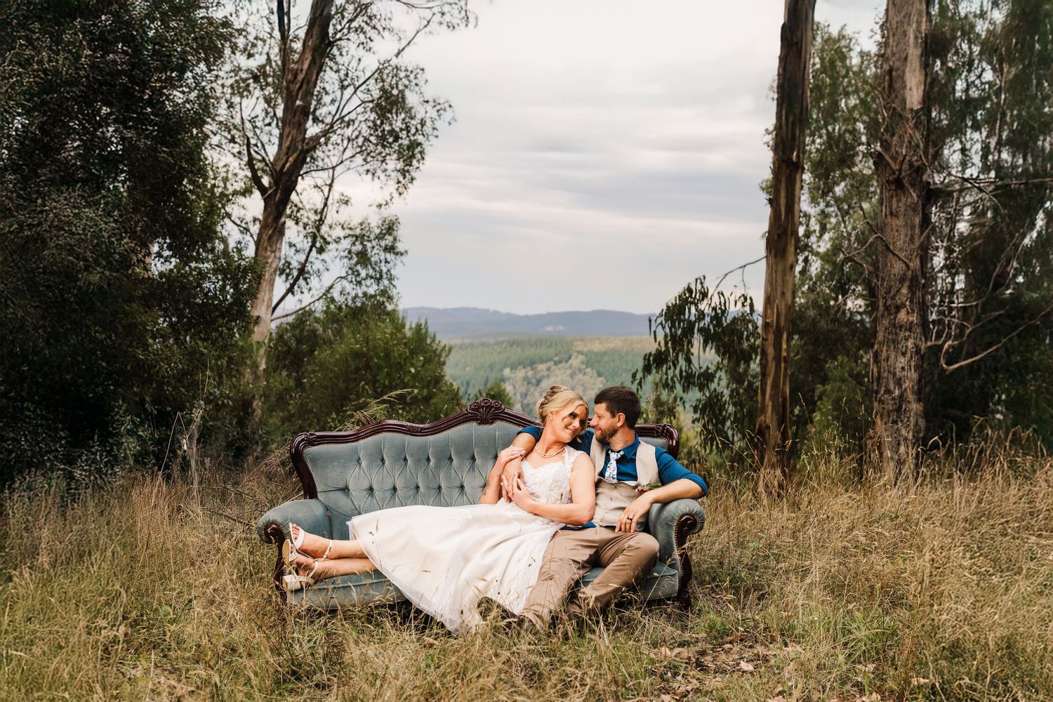 A couple in wedding attire relax together on a vintage blue sofa in a scenic outdoor field with mountain views.