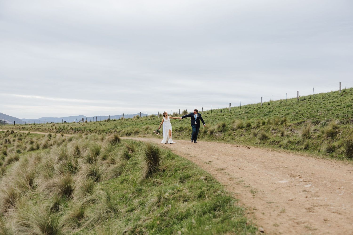 Two figures walk along a scenic dirt path through rolling green hills on an overcast day.