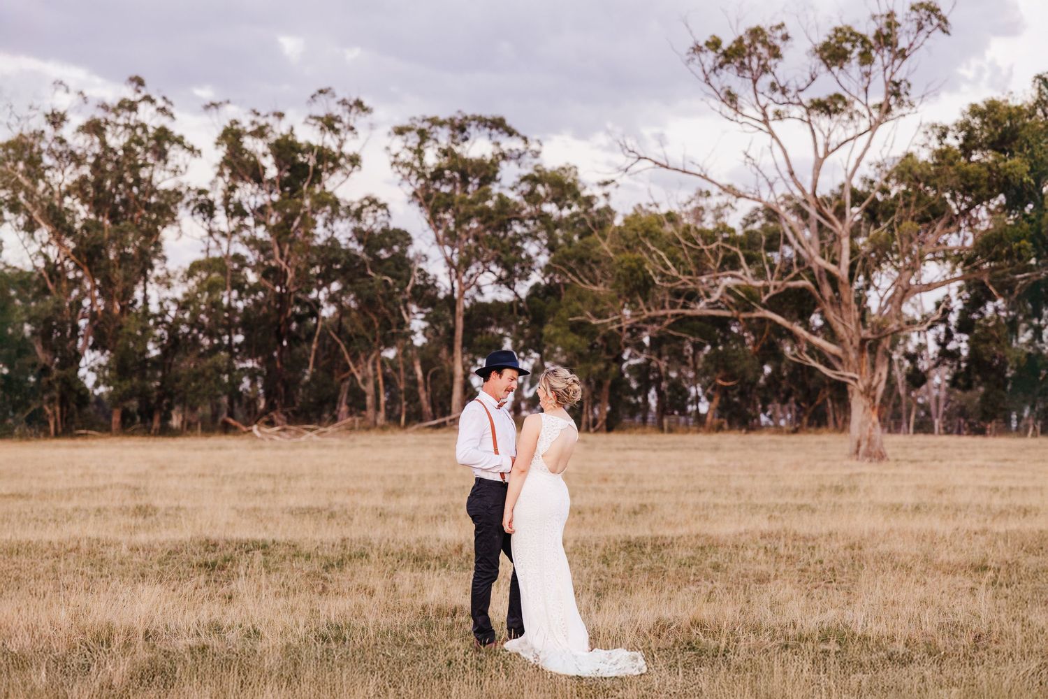 A couple embraces in their wedding attire on a rustic field with towering eucalyptus trees in the background.