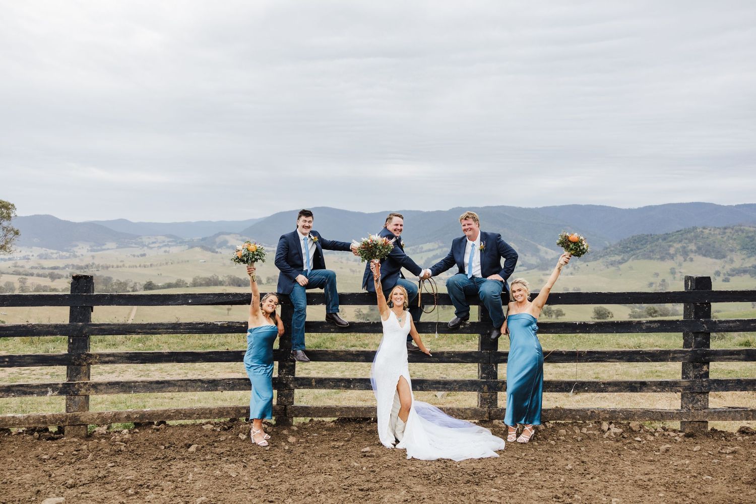 Wedding party members sitting casually on a rustic wooden fence with mountains in the background.