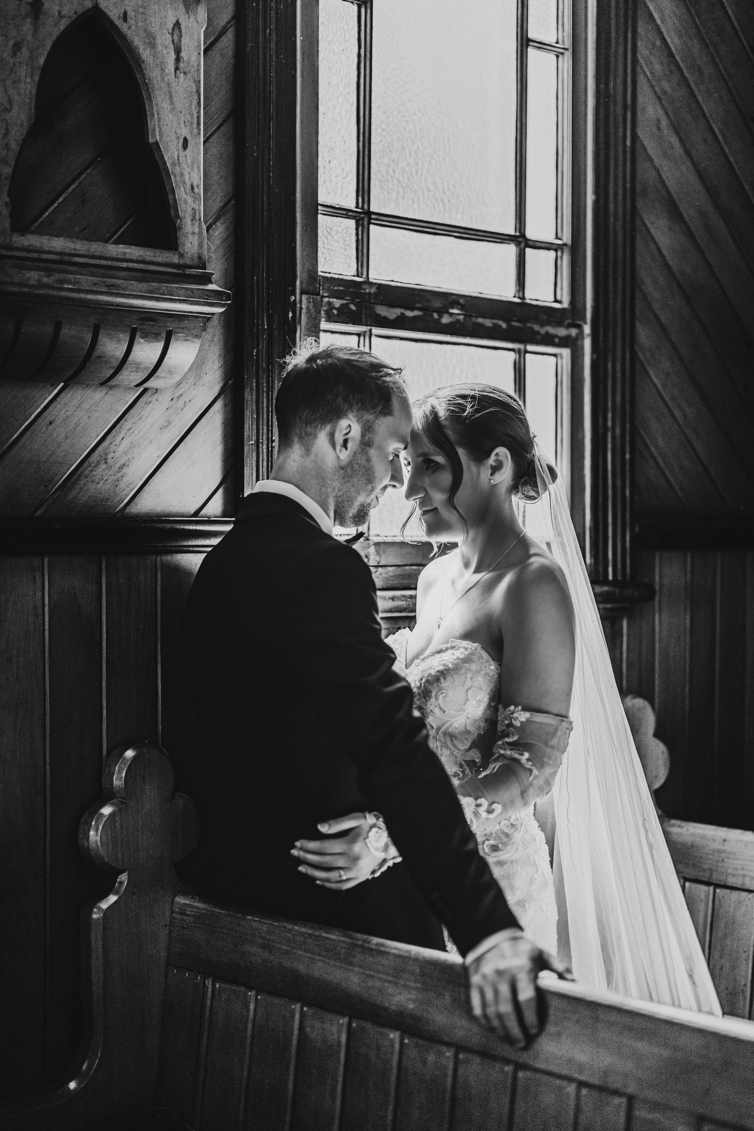 A romantic black and white moment between a wedding couple looking into each other's eyes by a rustic church window.