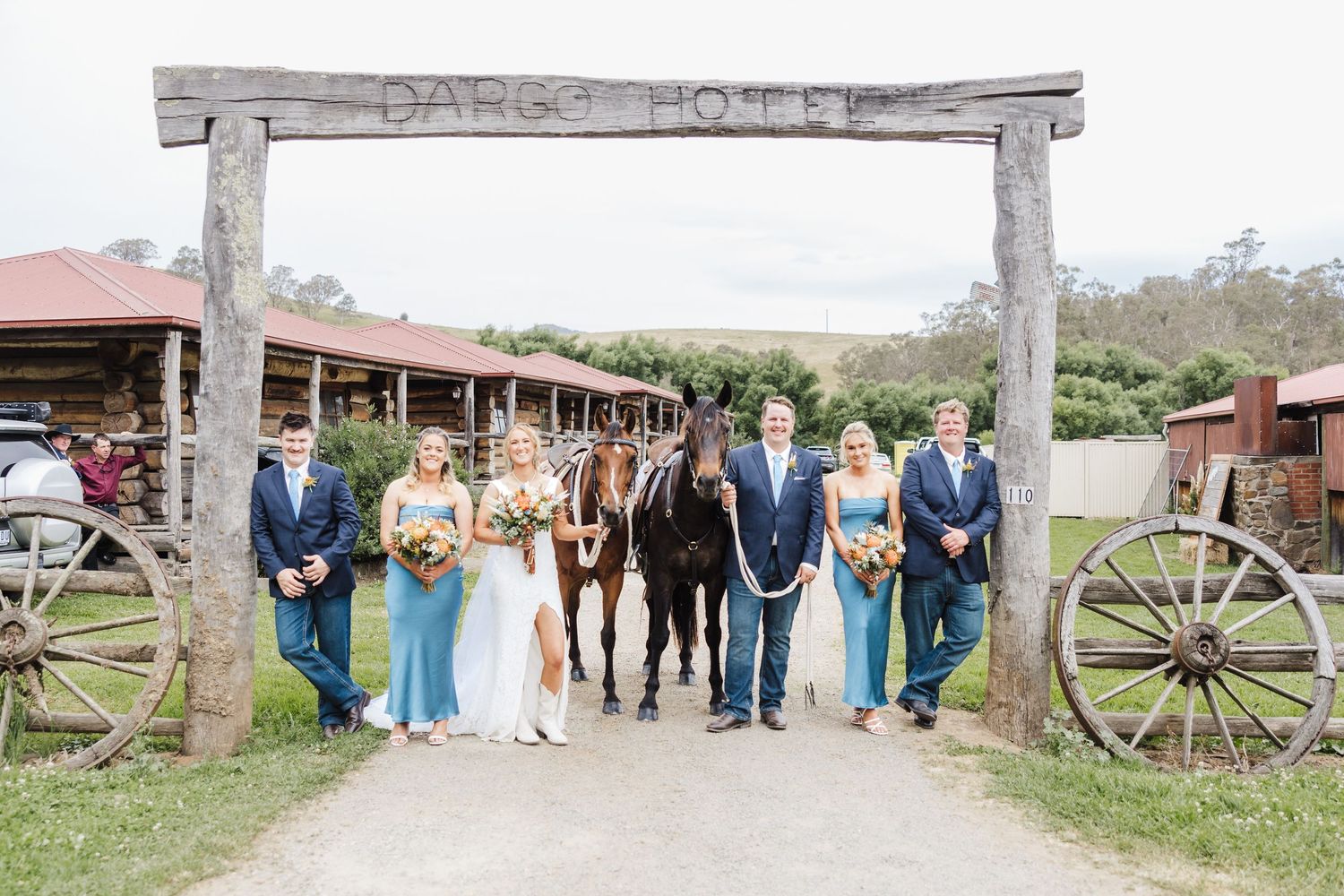 Wedding party poses beneath rustic wooden ranch entrance with wagon wheels and horse at country venue.