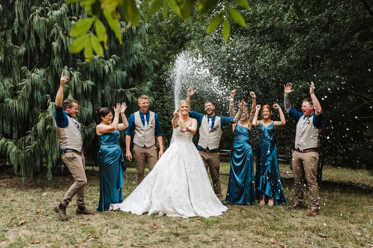 Wedding party celebrates outdoors with champagne spray while standing in front of lush green foliage.