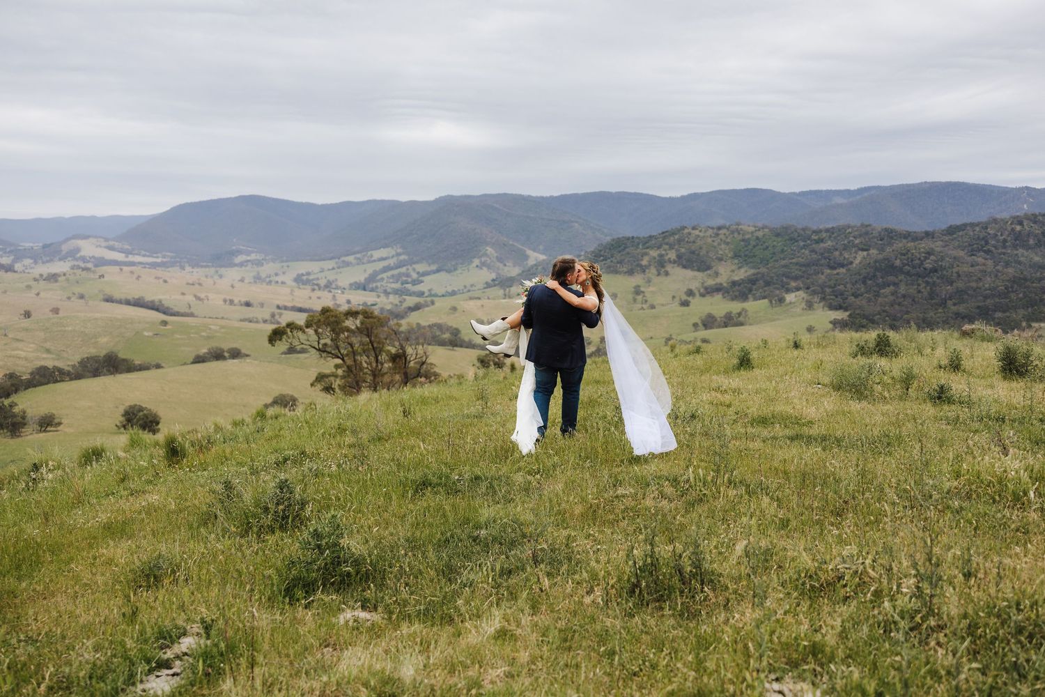 A newly married couple stands together on a grassy hilltop overlooking rolling mountains on a cloudy day.