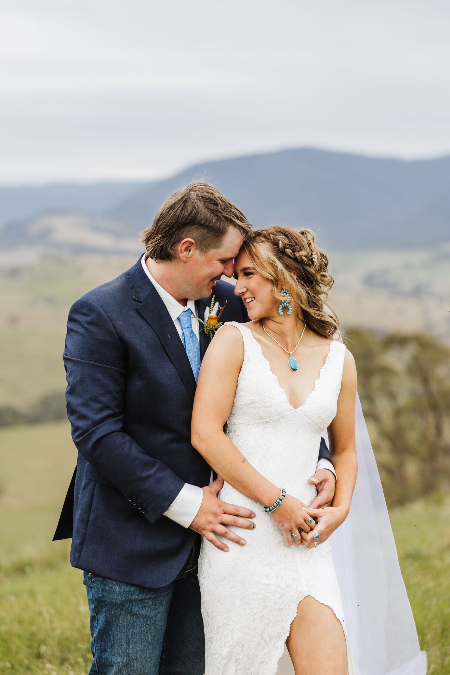 A romantic wedding couple shares an intimate moment on a hilltop with mountains in the background.