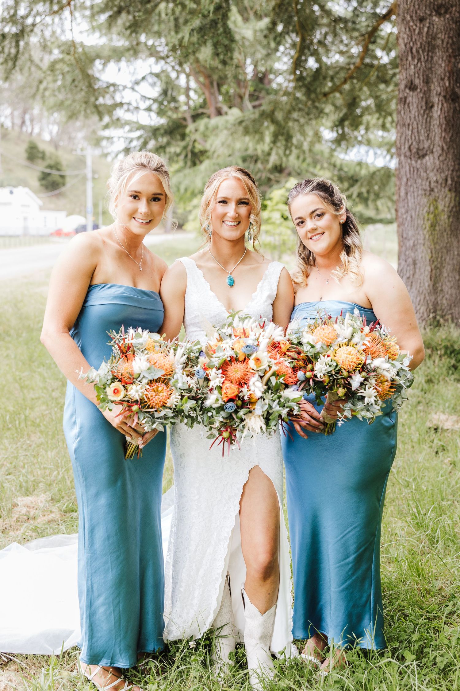Three women in formal attire stand together outdoors holding vibrant orange and white floral bouquets.