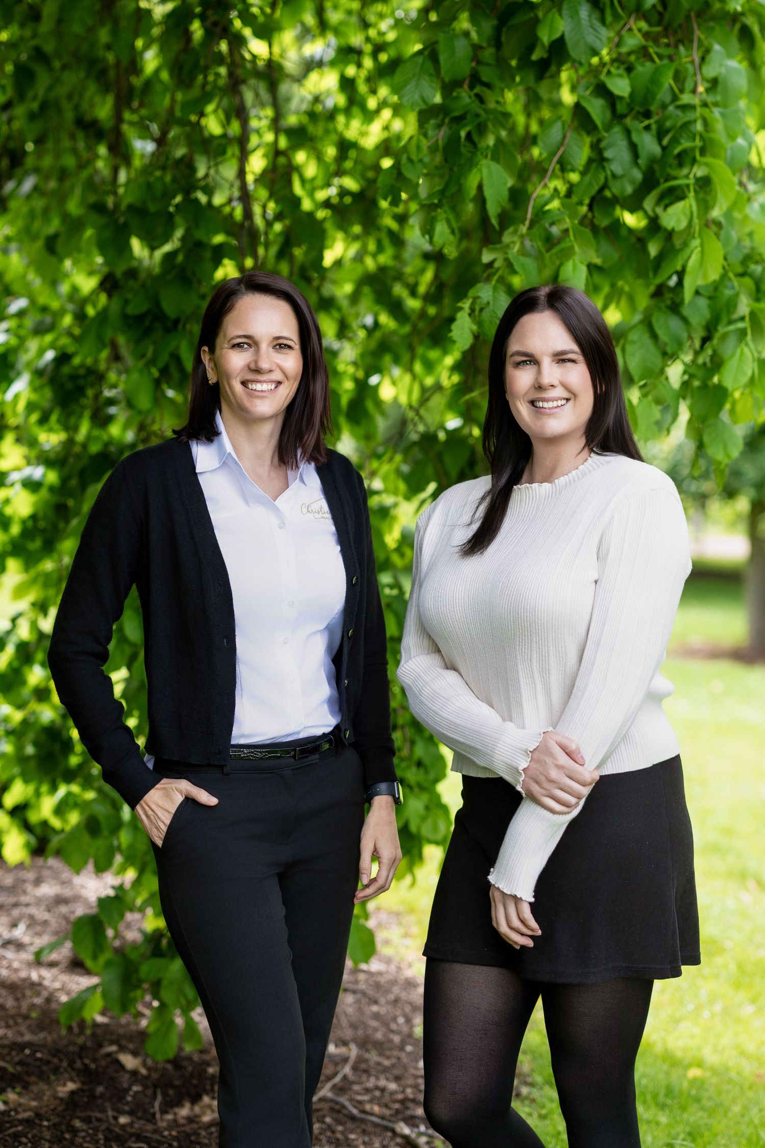 Two professionals in business attire pose outdoors against green foliage in a garden setting.