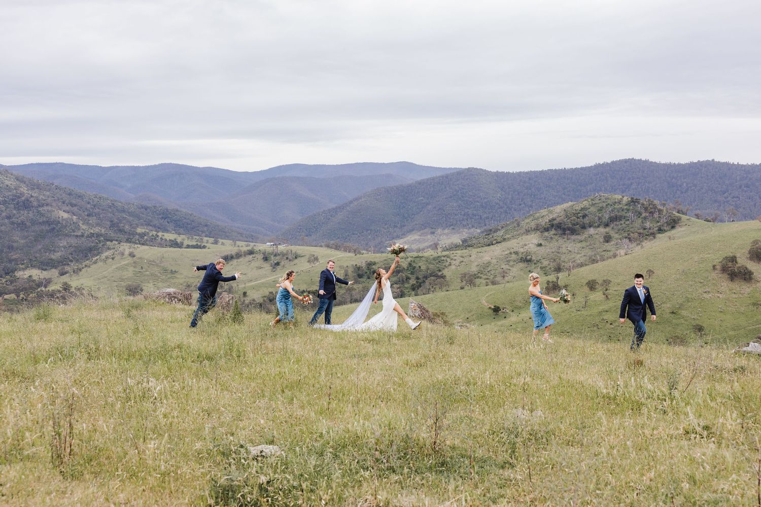 A group walks through a grassy mountain meadow with rolling hills under cloudy skies.