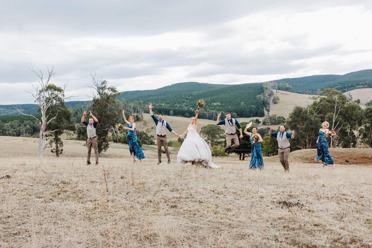 A wedding party jumps joyfully in unison against a backdrop of rolling hills and mountains on a cloudy day.