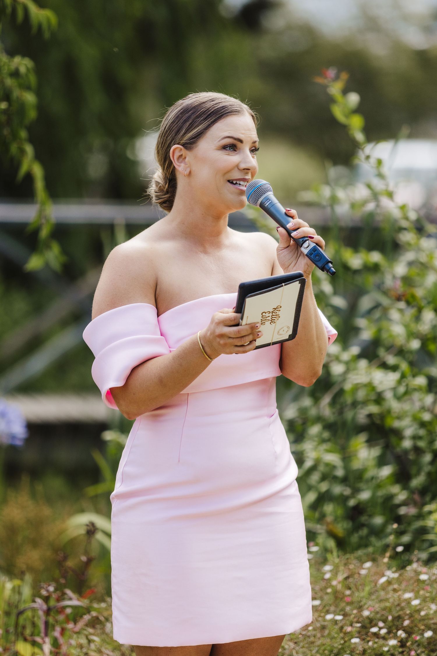 A person in a pink off-shoulder dress delivers an emotional speech while holding a book and microphone outdoors.