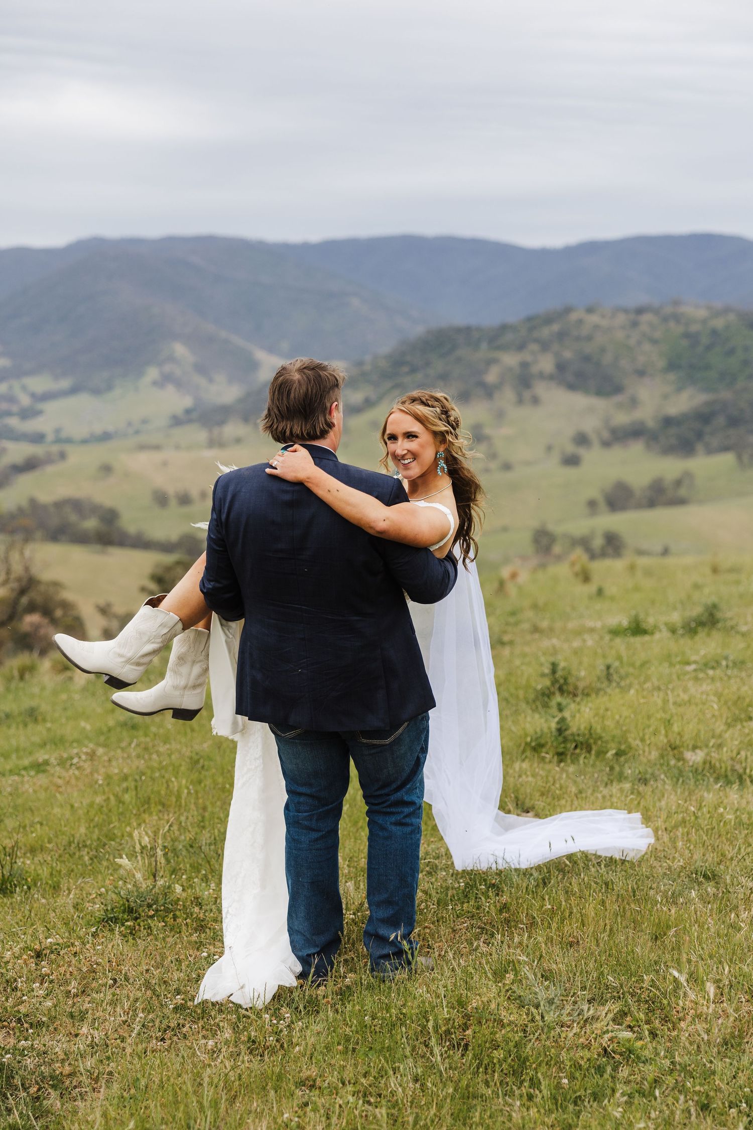 A romantic wedding couple embraces on a green hillside with rolling mountains in the background on their special day.