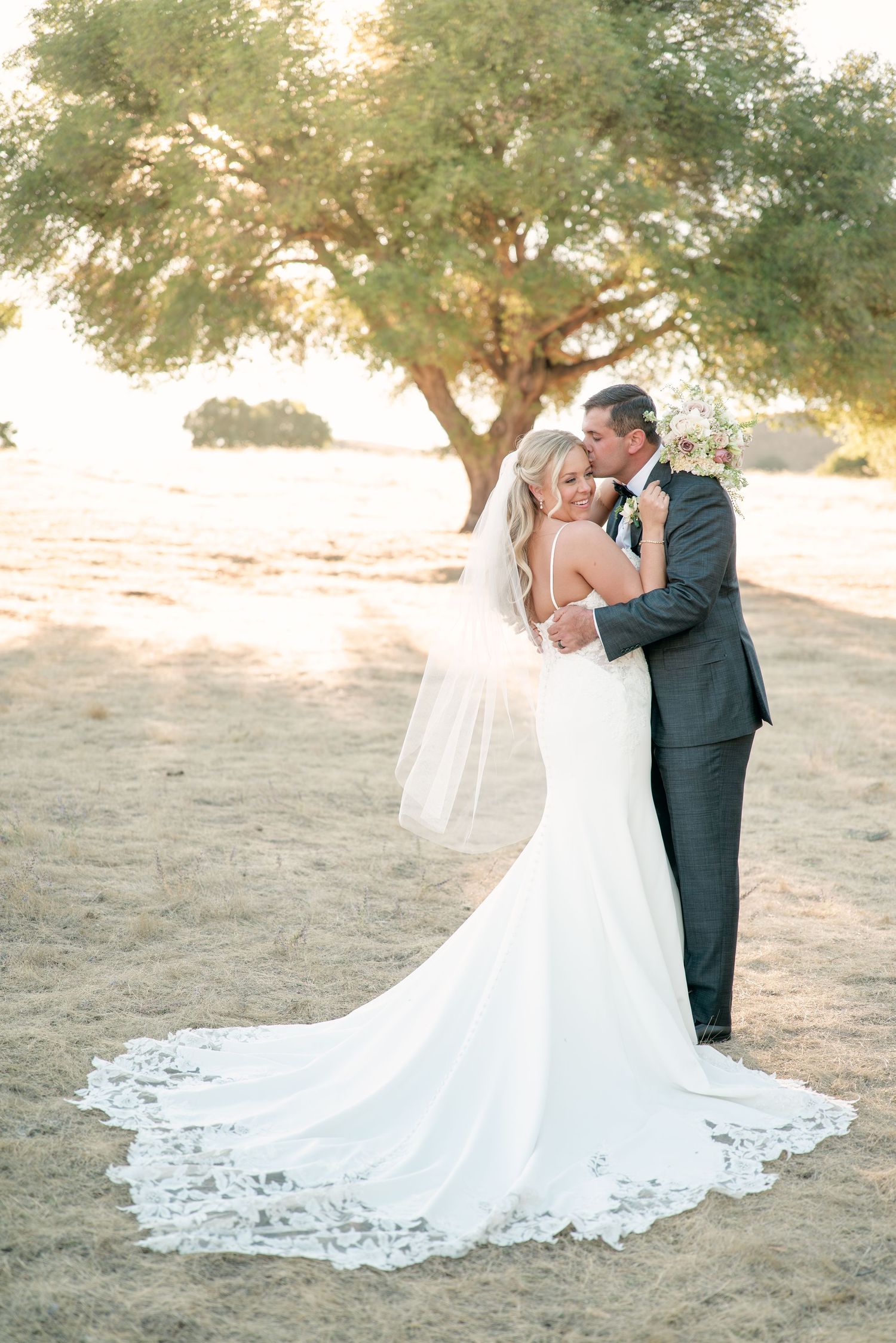 A romantic wedding couple embraces under a majestic tree at golden hour, with the bride's lace gown trailing behind.