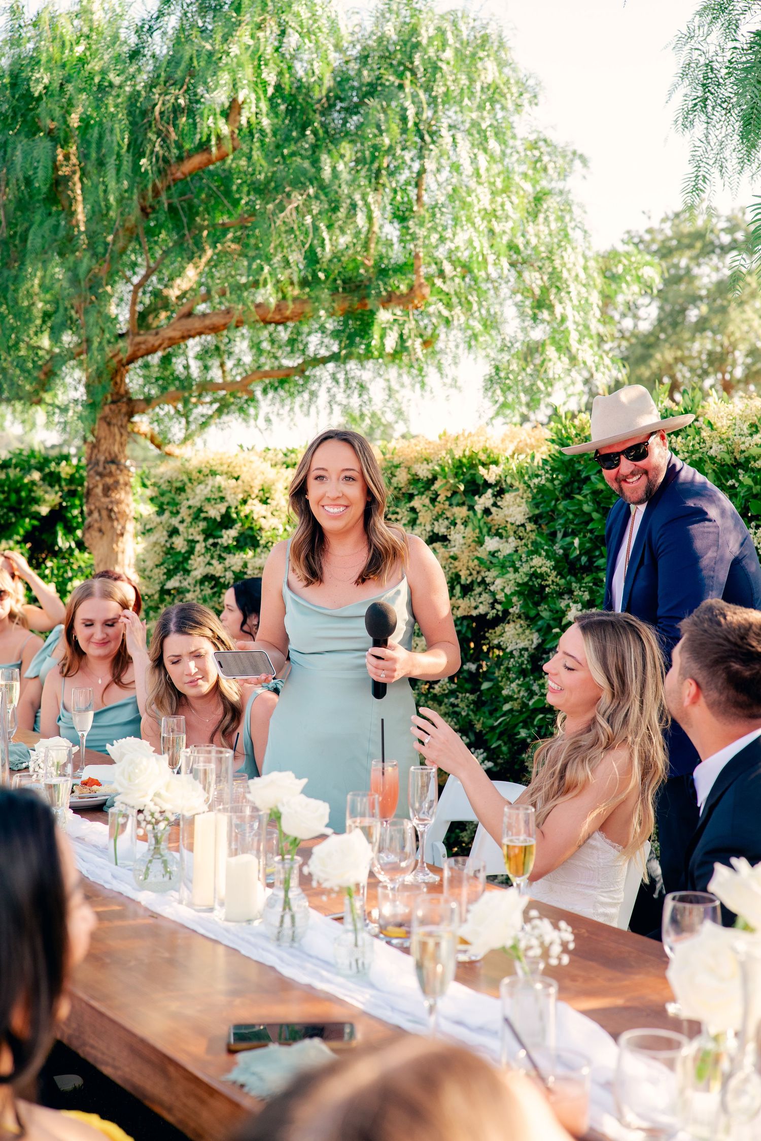 Guests enjoy an outdoor wedding reception dinner at a long table under a willow tree with glowing candle centerpieces.