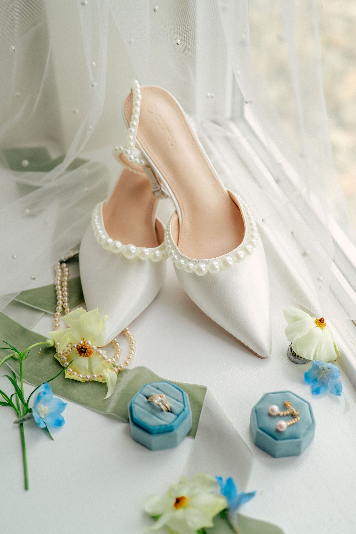 Elegant white bridal shoes with pearl trim displayed alongside blue hexagonal ring boxes and delicate yellow and blue flowers.