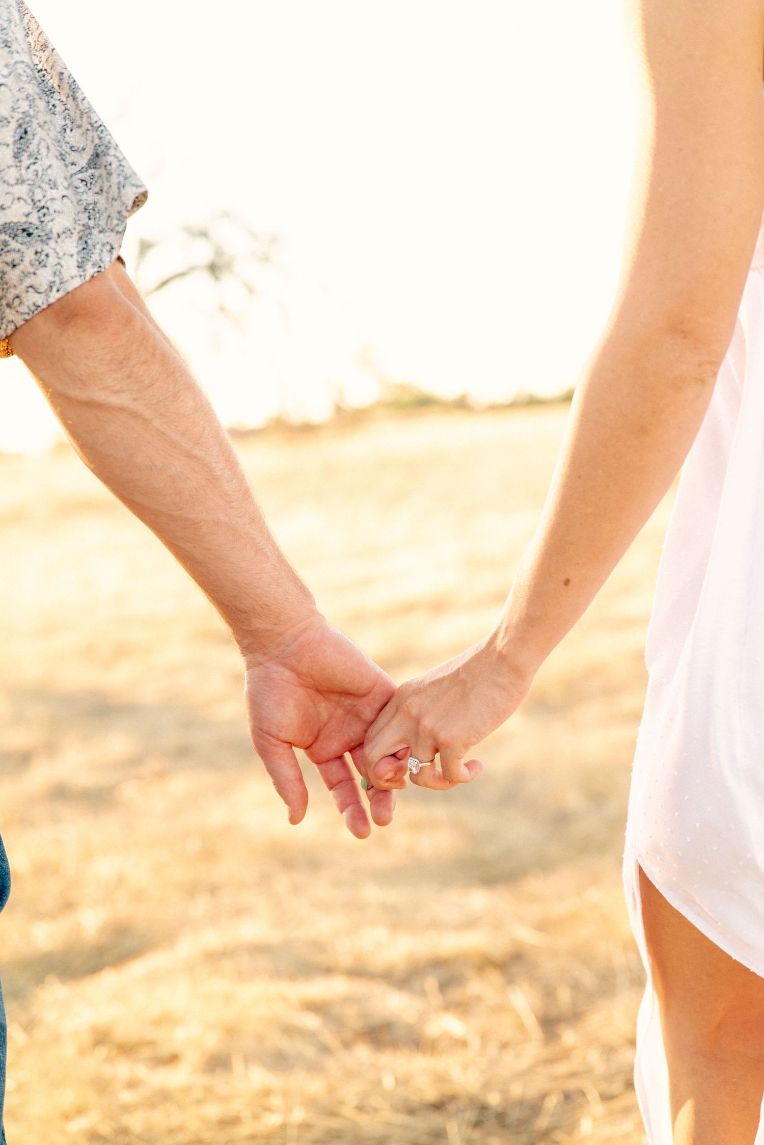 Close-up of two people holding hands at sunset with a golden field in the background.