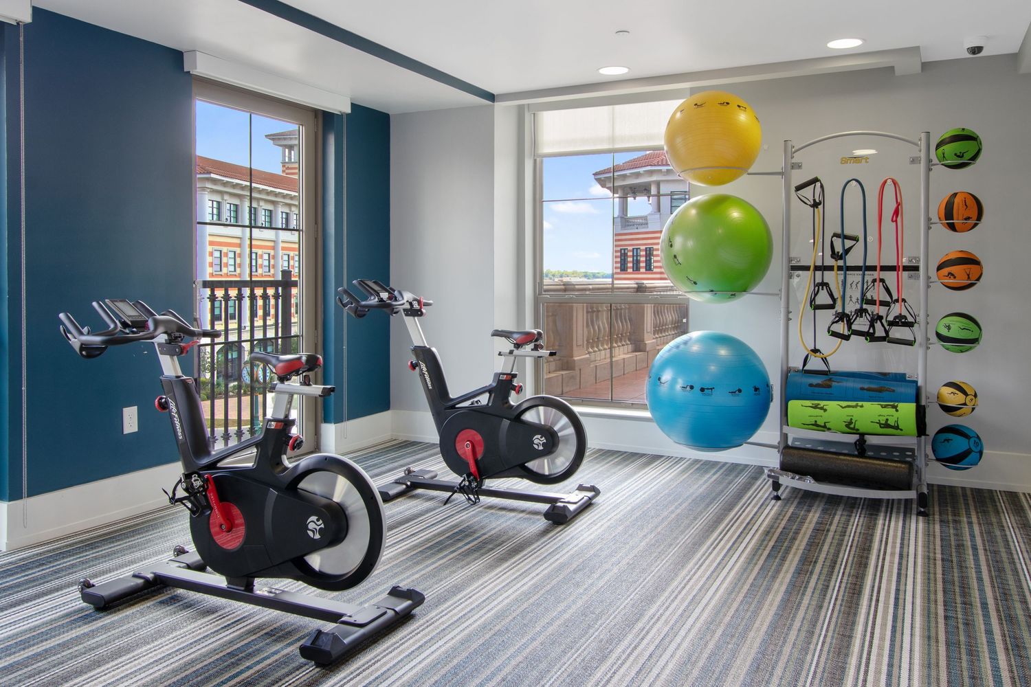 Modern home gym room with exercise bikes, stability balls in various colors, and fitness equipment against blue accent wall.