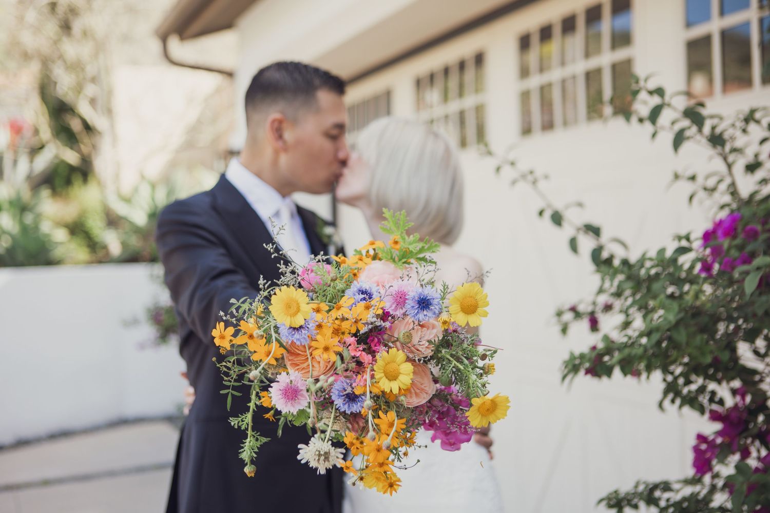 A wedding couple poses in front of a building while holding a vibrant bouquet of mixed flowers.