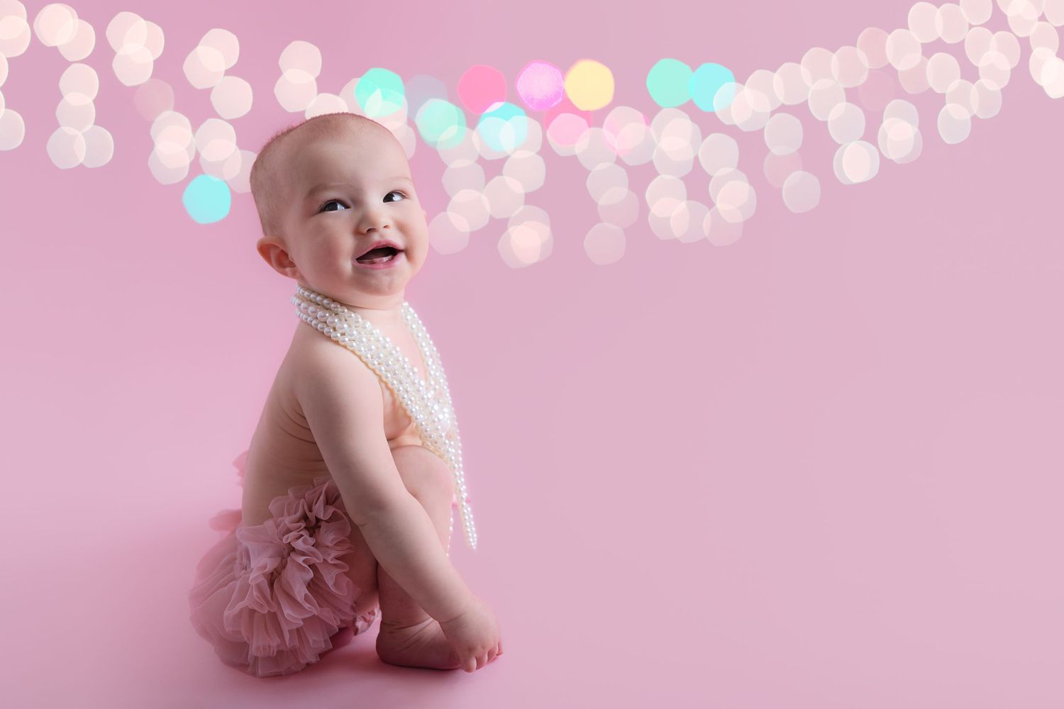 Baby wearing a pink tutu sits on pink background with string lights and floral decorations.