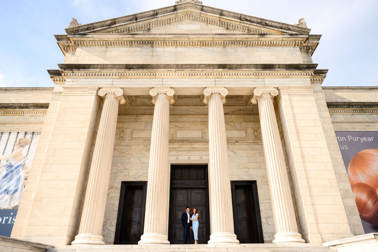 Two people stand at the entrance of a grand neoclassical building with tall columns and stone steps.