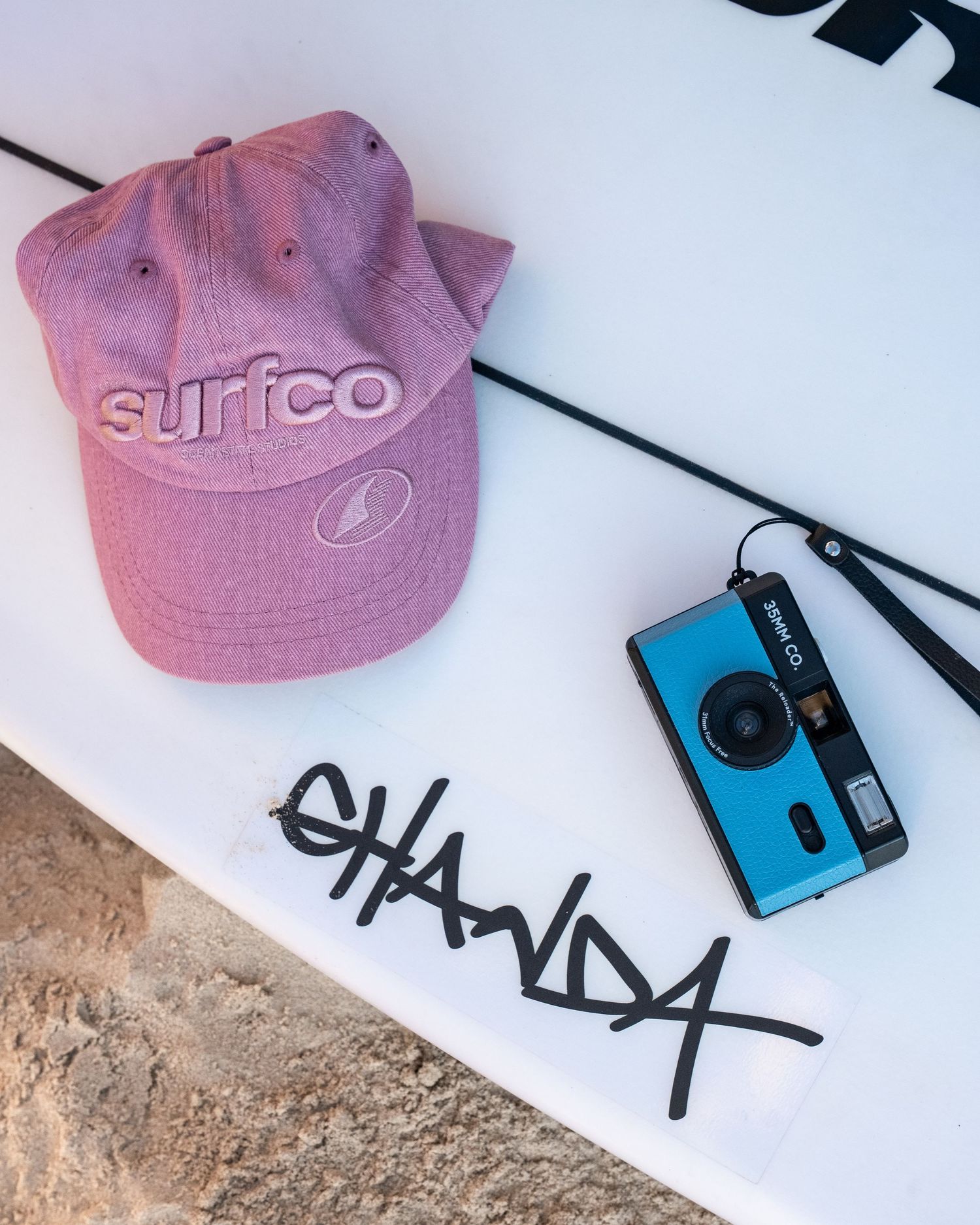 Pink Surfco cap and blue film camera resting on a white surfboard with Chanda graffiti signature.