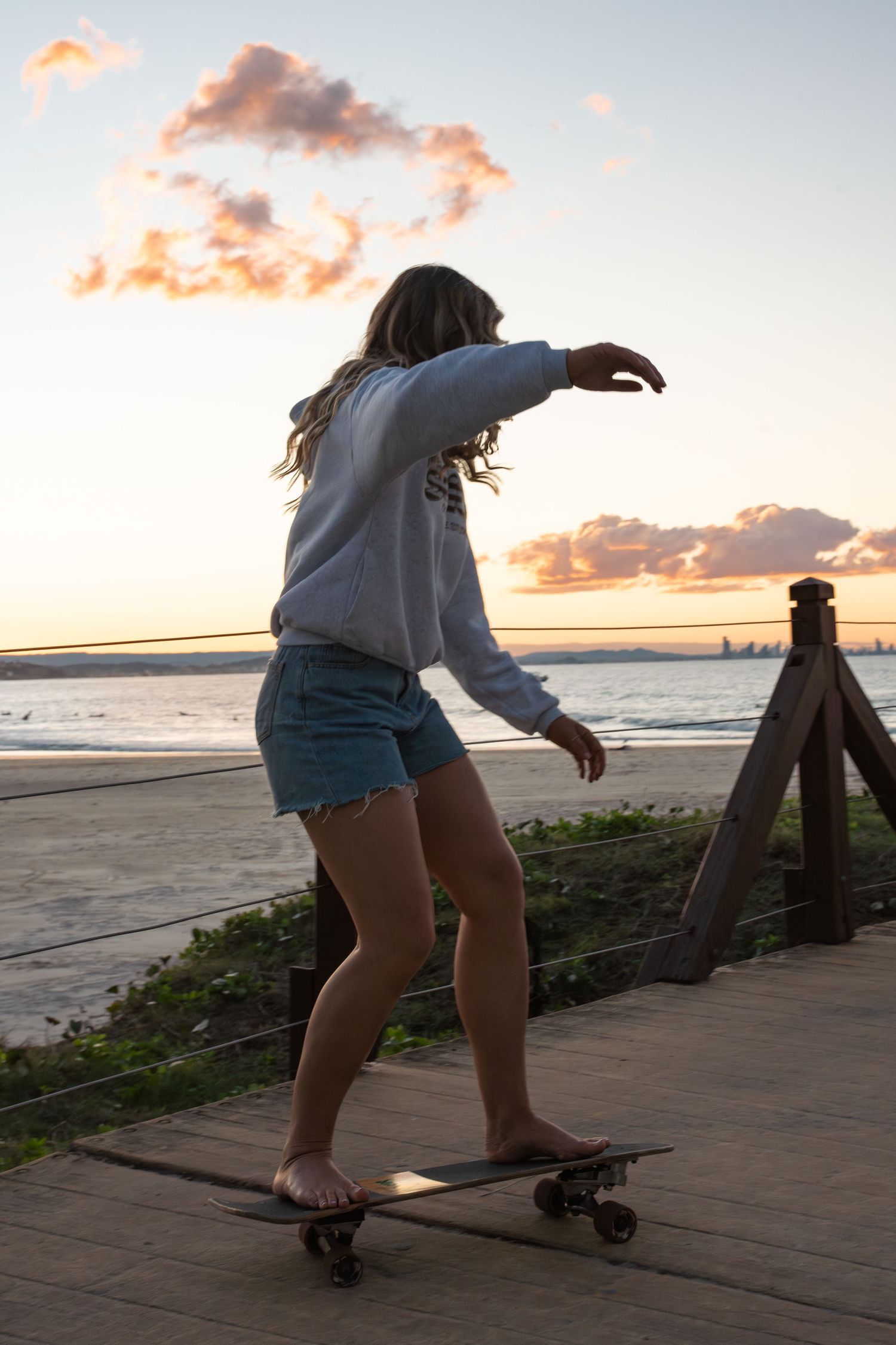 Young woman skateboarding barefoot on a beachside boardwalk at sunset, wearing denim shorts and a gray hoodie.