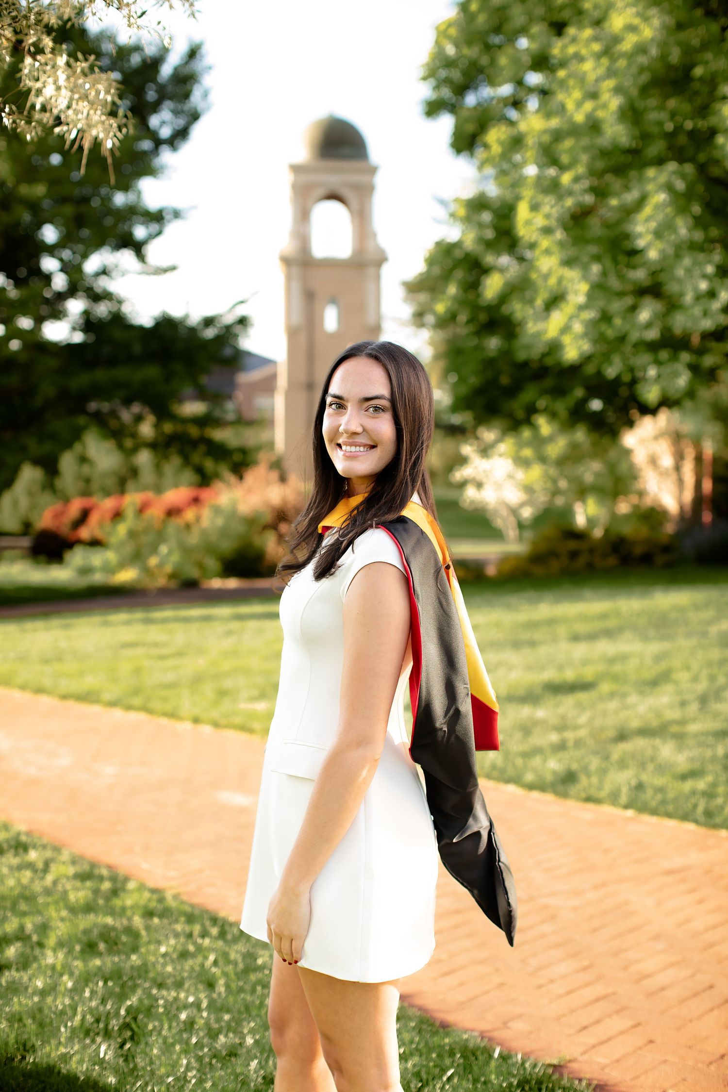 Student in white dress standing on brick path with bell tower and green trees in background during golden hour.