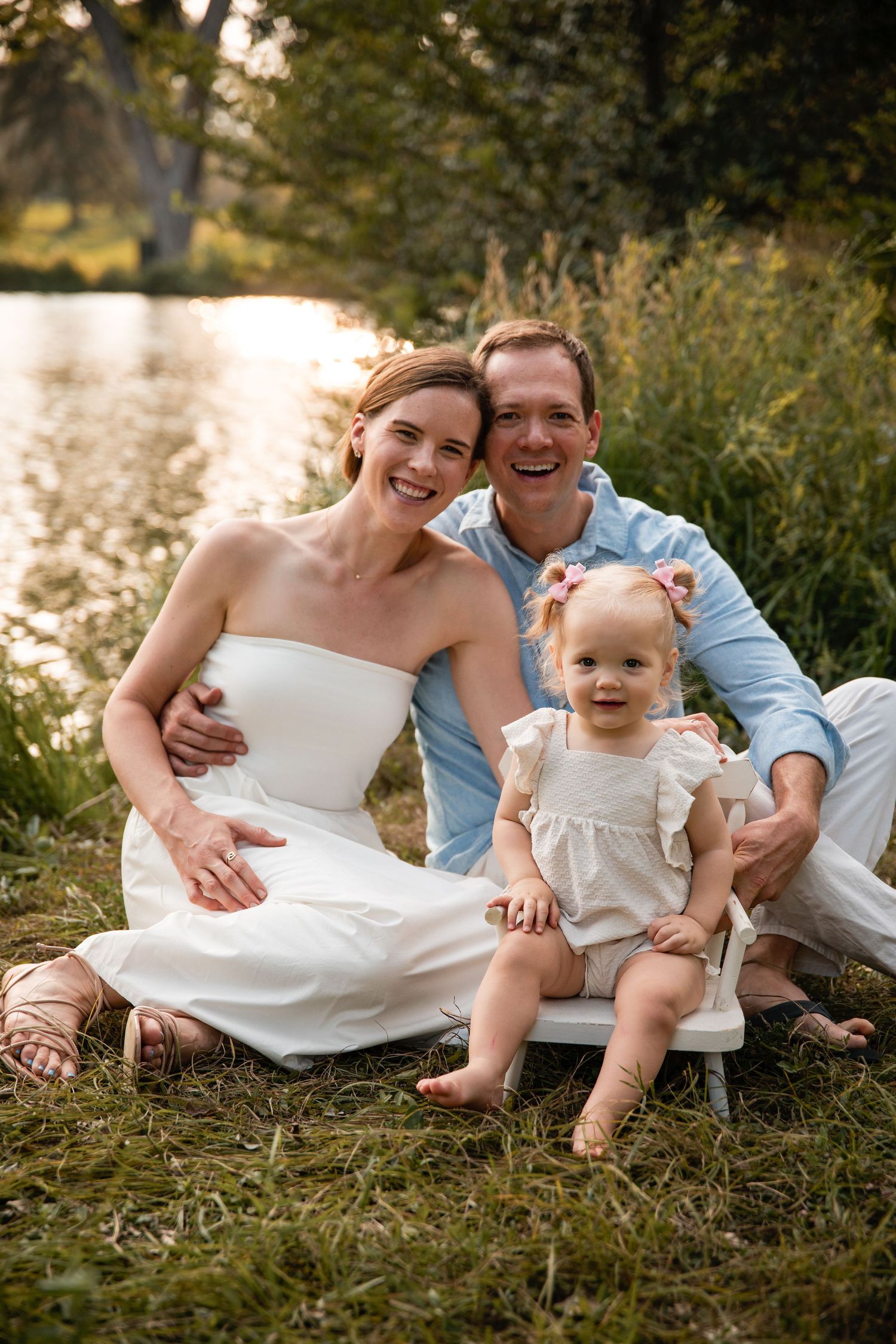Happy family in white outfits sitting together by a peaceful lake during golden hour sunset.