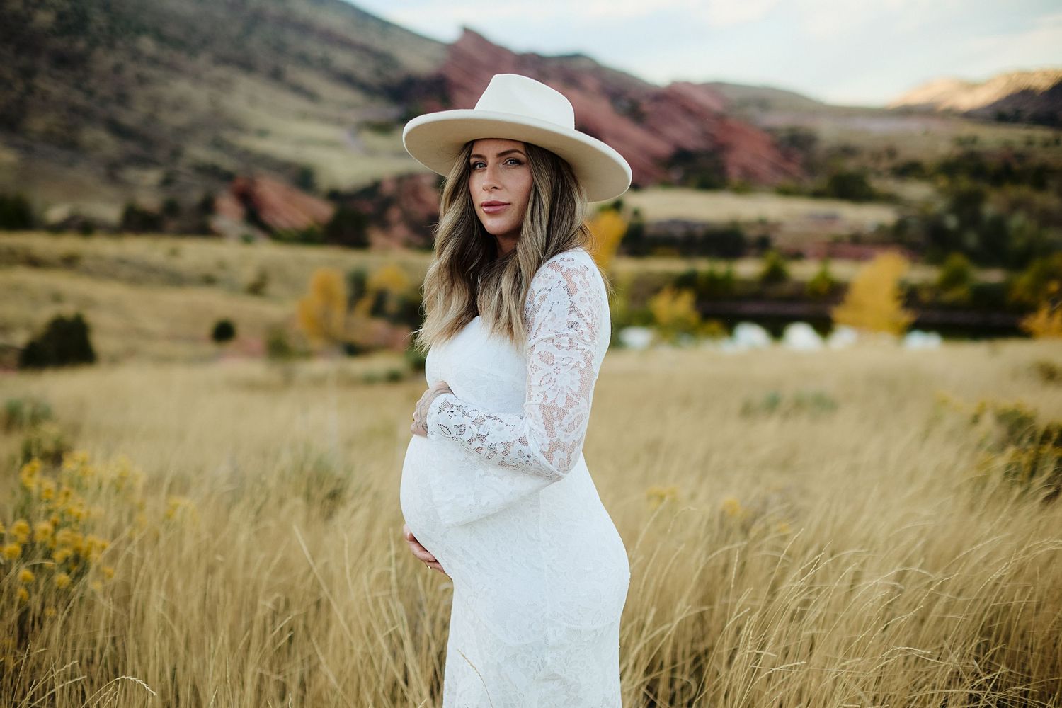 A woman in a white lace dress and wide-brimmed hat stands in a golden wheat field at sunset.