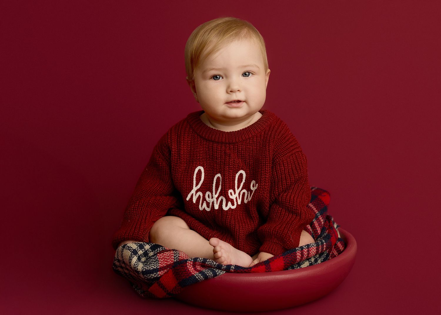 A baby wearing a red holiday sweater sits in a red bowl against a burgundy background.