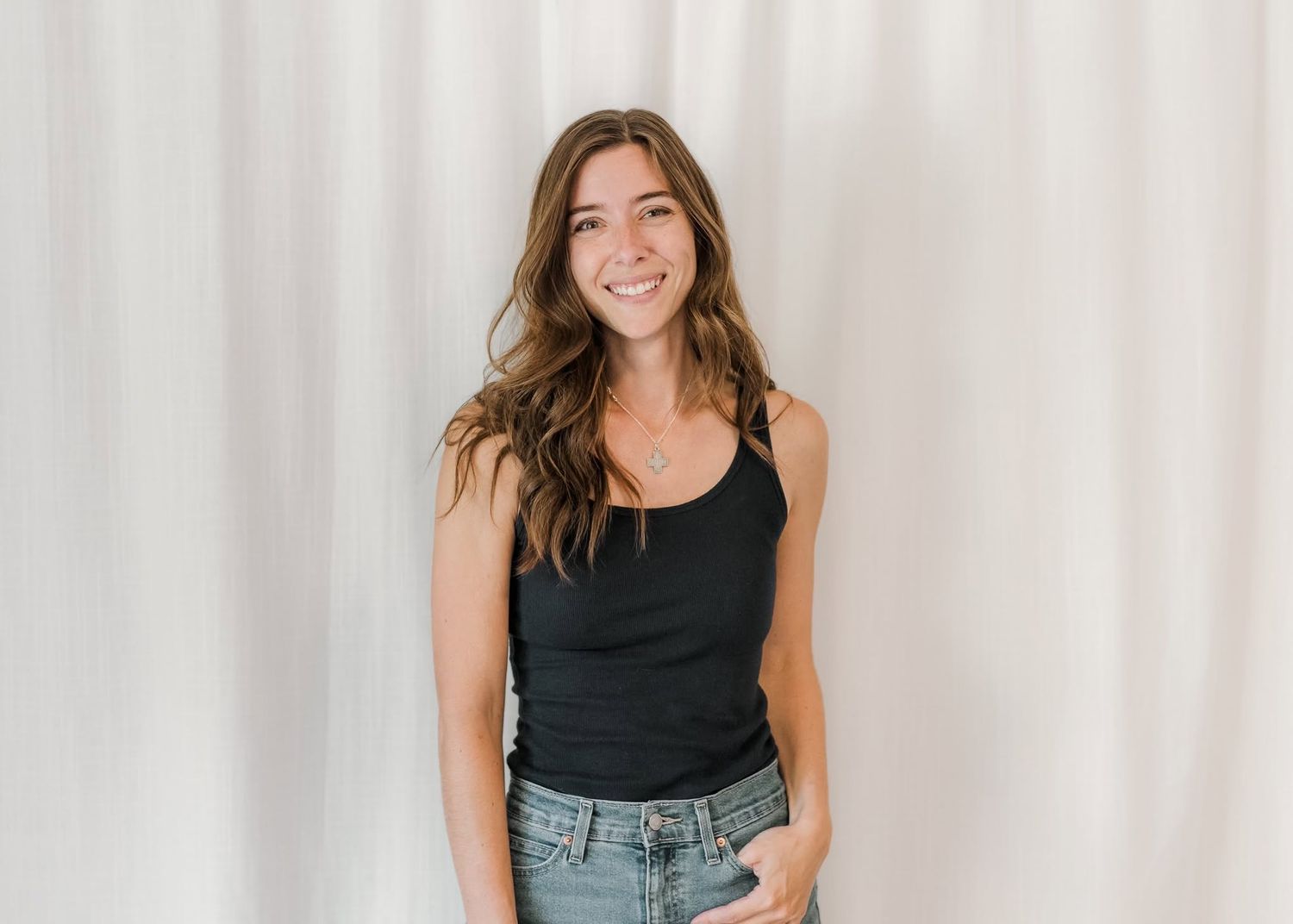 Smiling young woman with long brown hair wearing a black tank top and jeans, posing against a white curtain backdrop.