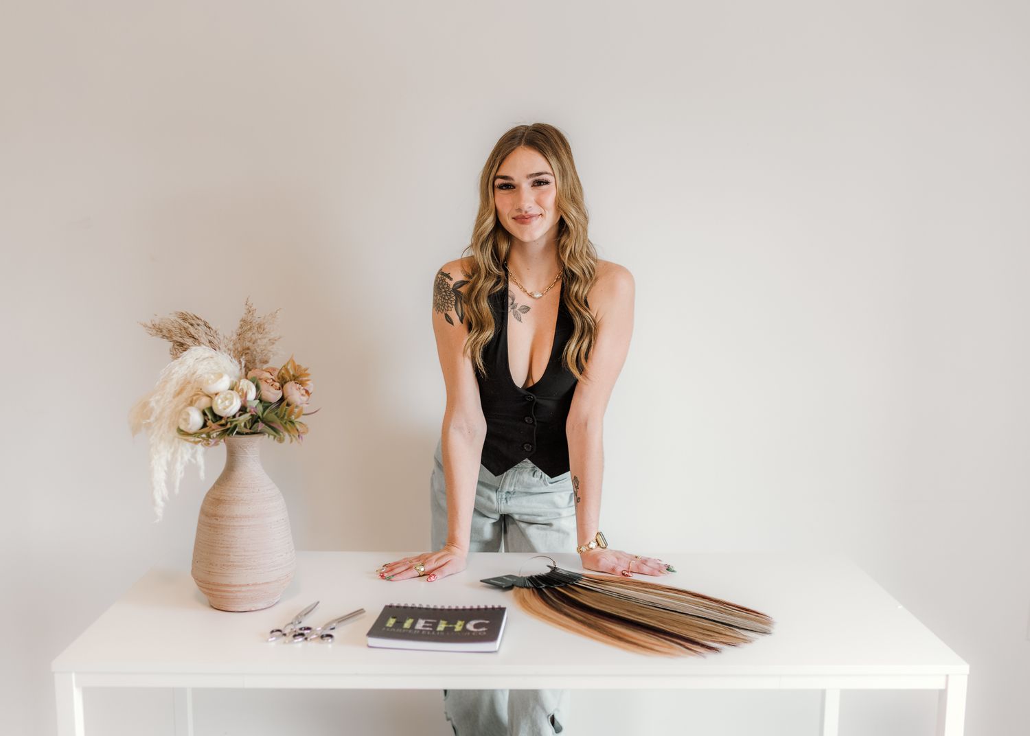 Woman standing at white desk with hair extensions, flowers, and branded materials in a bright studio setting.