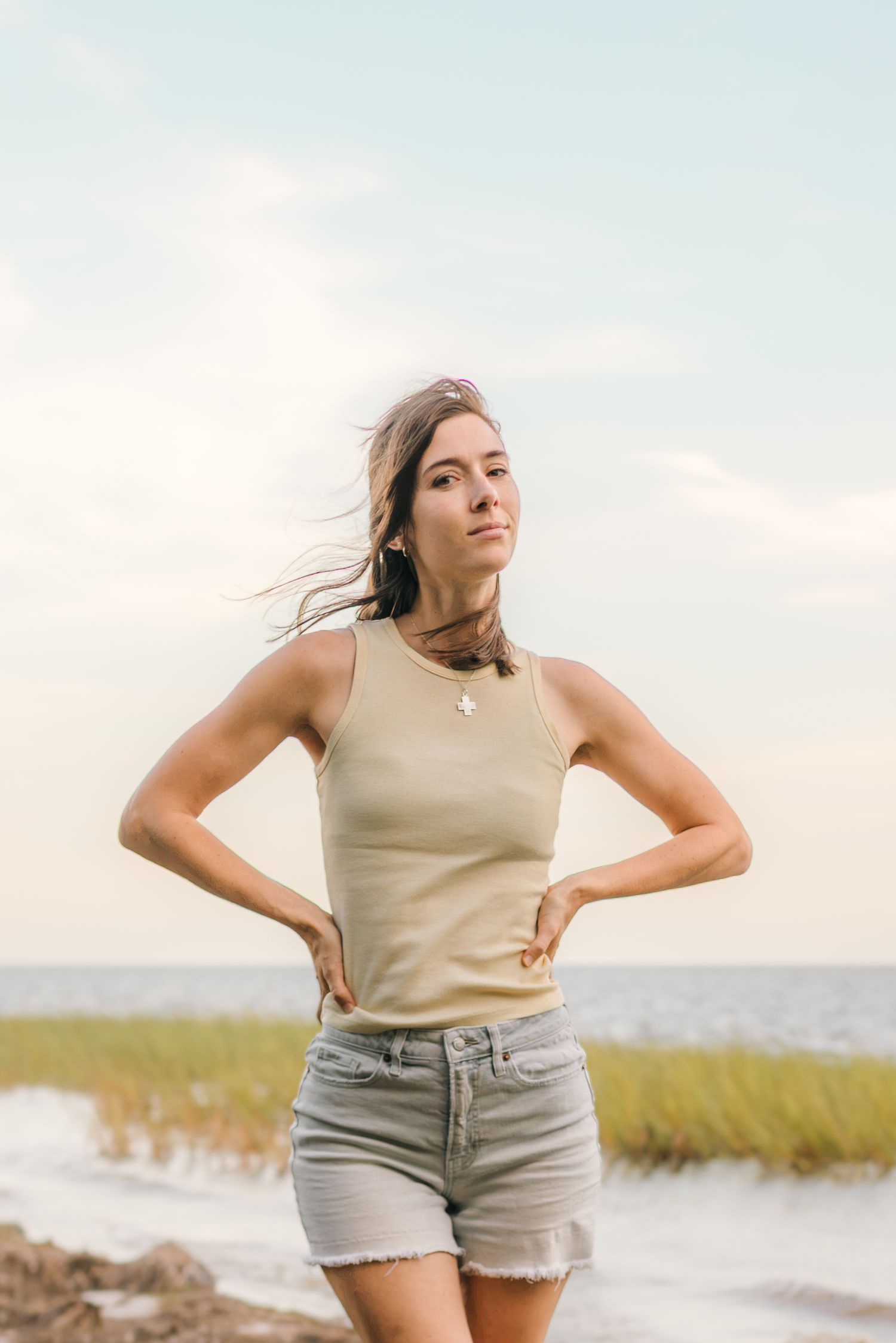Young woman in beige tank top and denim shorts poses confidently on a coastal beach with marsh grass and ocean backdrop.