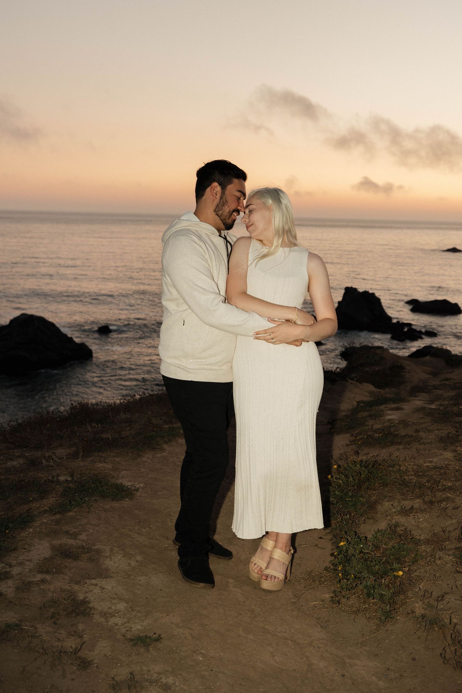 A romantic couple embraces on a coastal cliff at sunset with the ocean and rocky shore in the background. Sonoma Coast Beach.