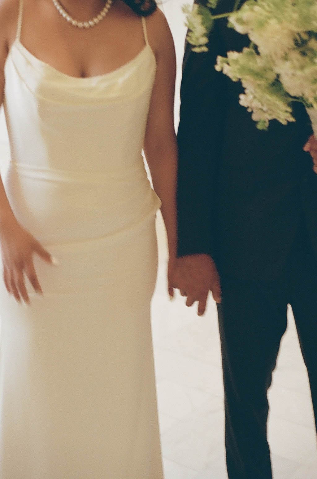 A couple in wedding attire holds hands, with the bride wearing a white satin gown and pearl necklace.