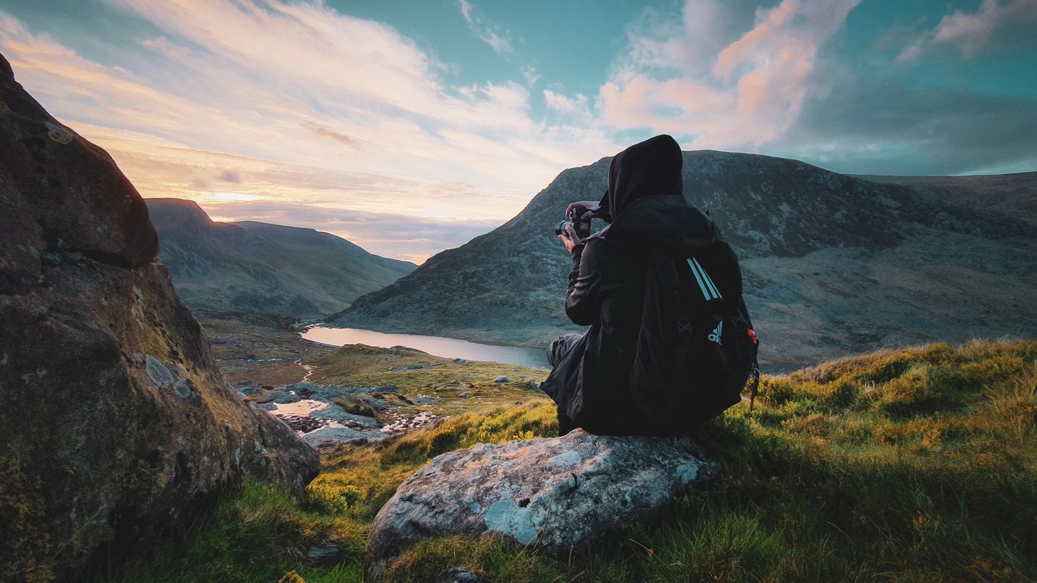 A figure sits on a rocky outcrop overlooking misty mountains and lakes during sunset in a dramatic landscape.