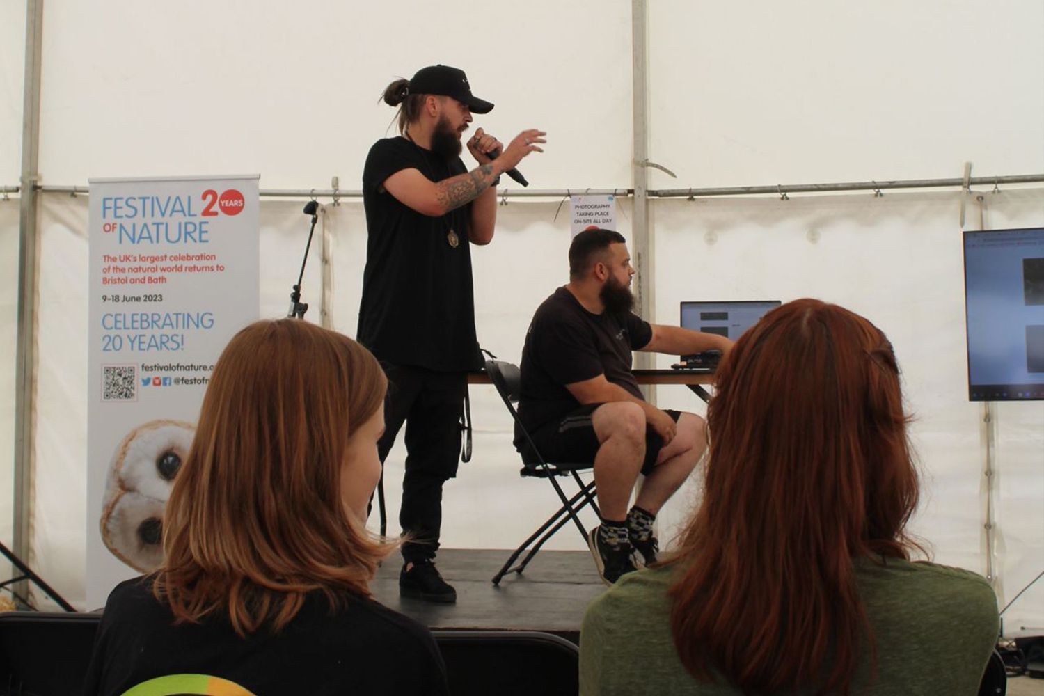People gathered in a white tent for an event presentation with speakers and audience members seated and standing.