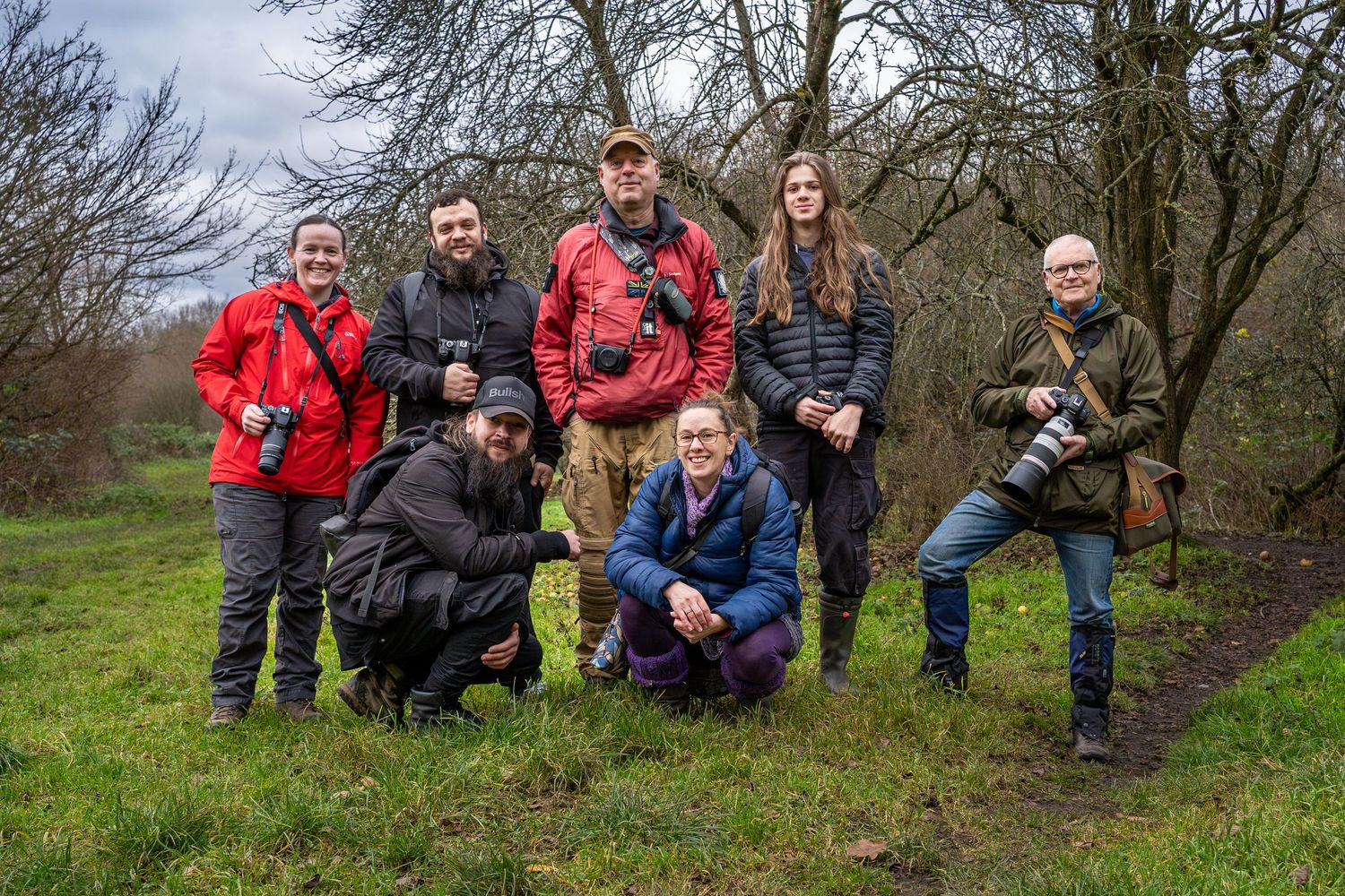 A group of people sitting and crouching on a grassy slope wearing outdoor gear and coats for hiking.