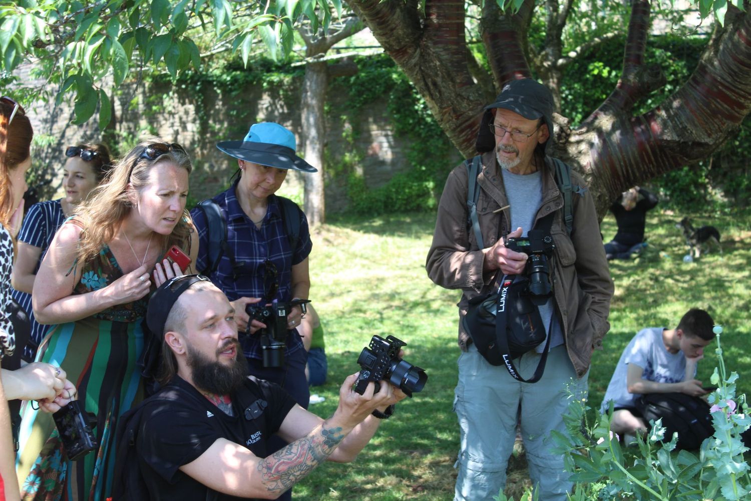 Group of photographers with professional cameras gather outdoors under shaded trees in a park setting.