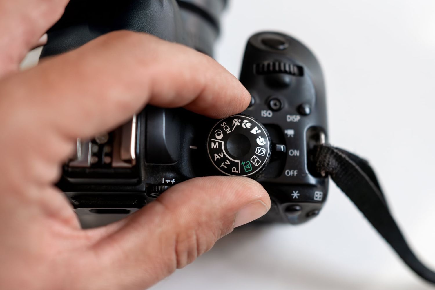 A hand adjusts the mode dial on a black DSLR camera, showing camera controls in close-up detail.