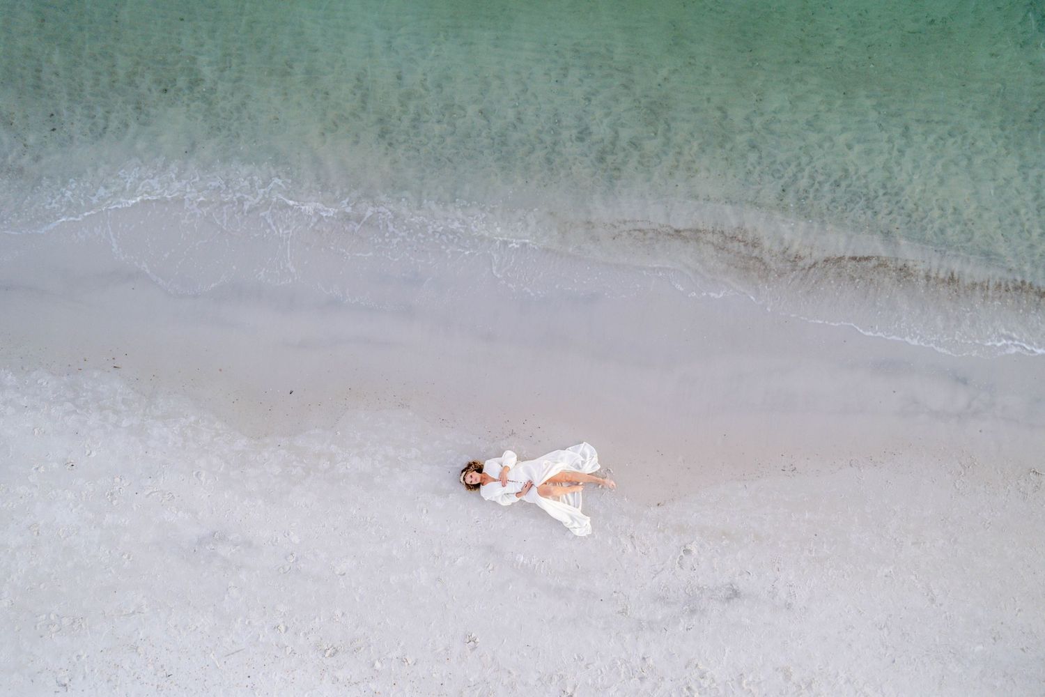 Aerial view of person in white dress lying on sandy beach next to turquoise waters.