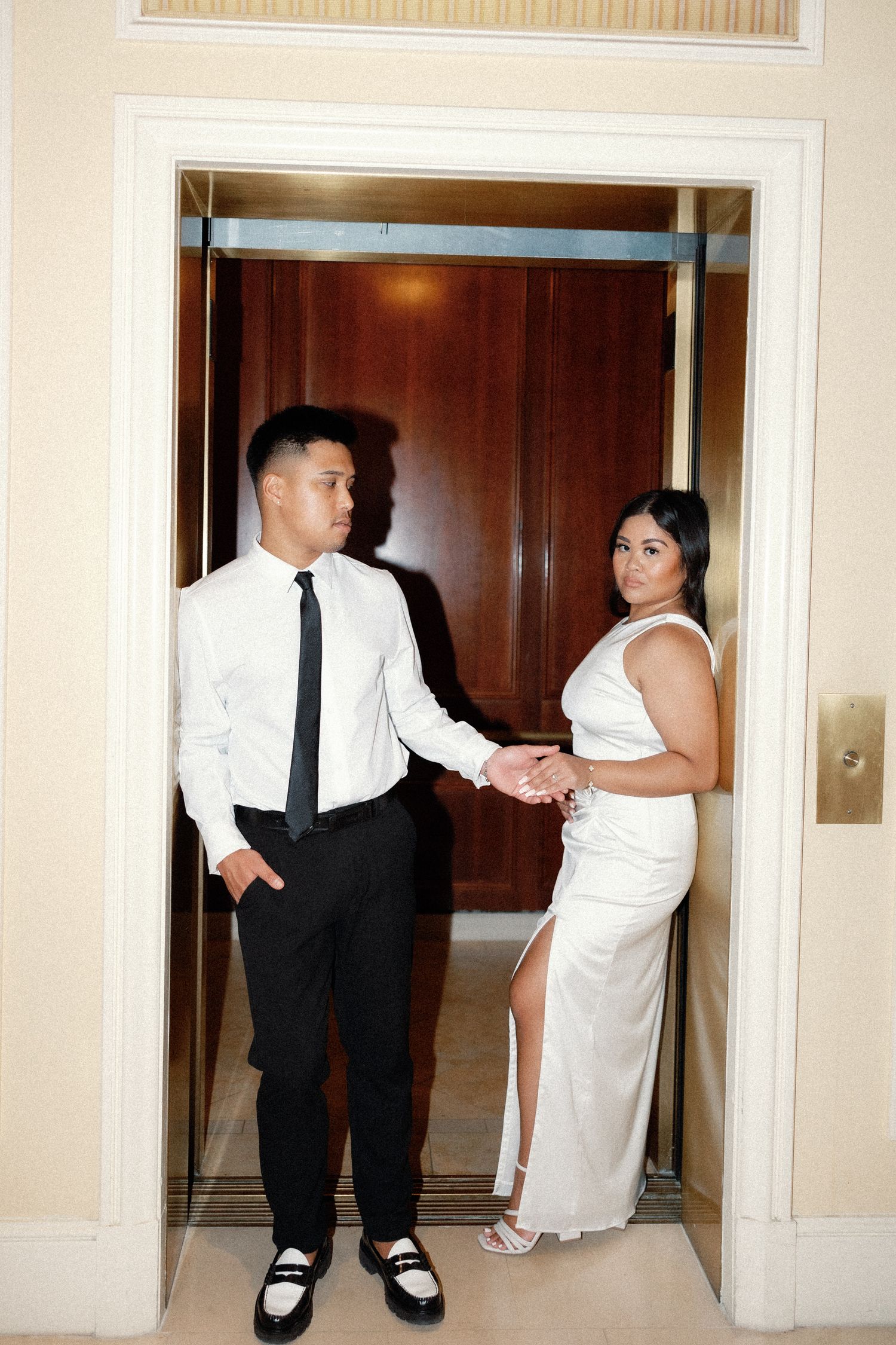 Elegant couple posing in elevator doorway, man in white shirt and black tie, woman in white slit gown holding hands.