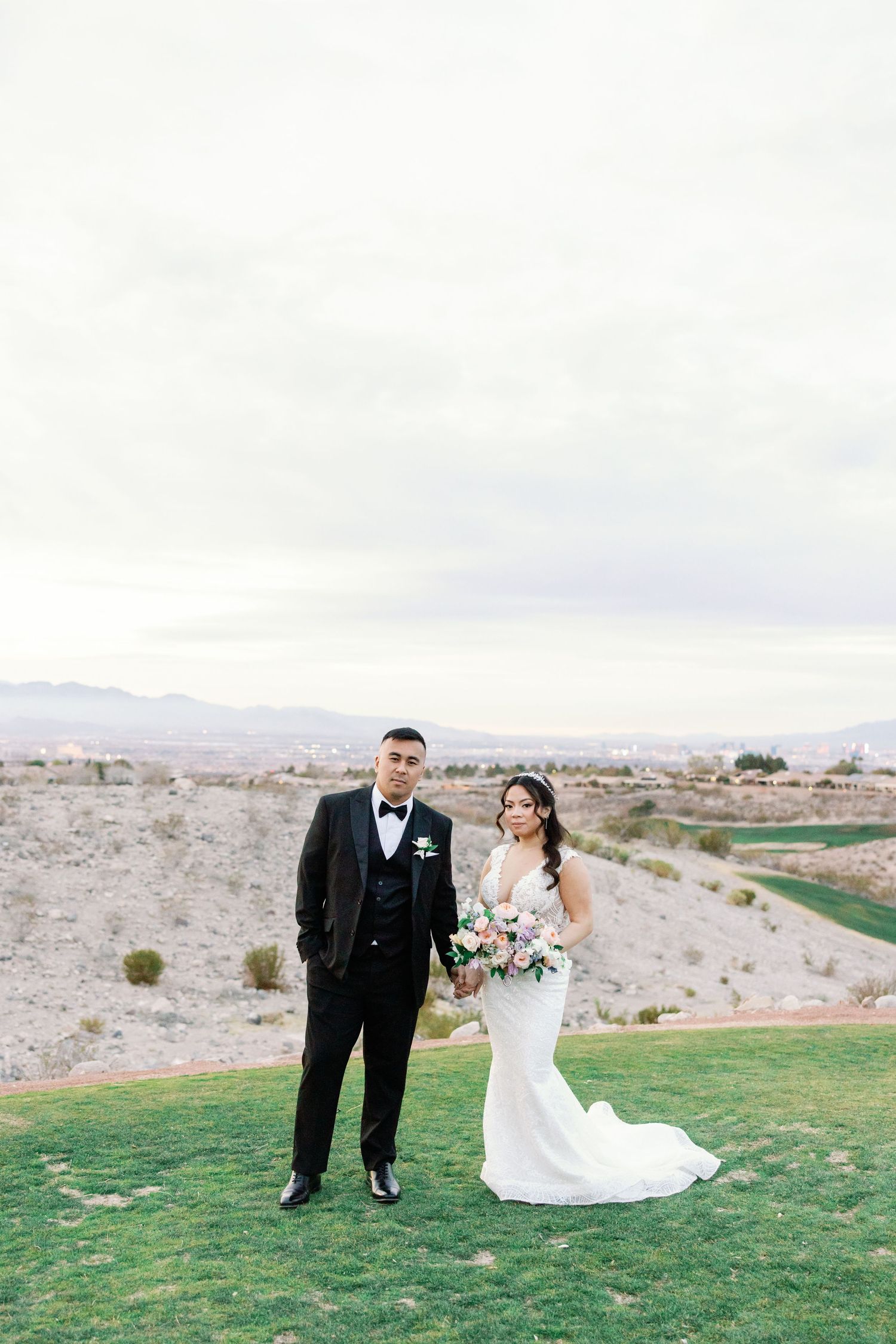 Bride and groom posing on a green hillside with a scenic desert landscape backdrop on their wedding day.