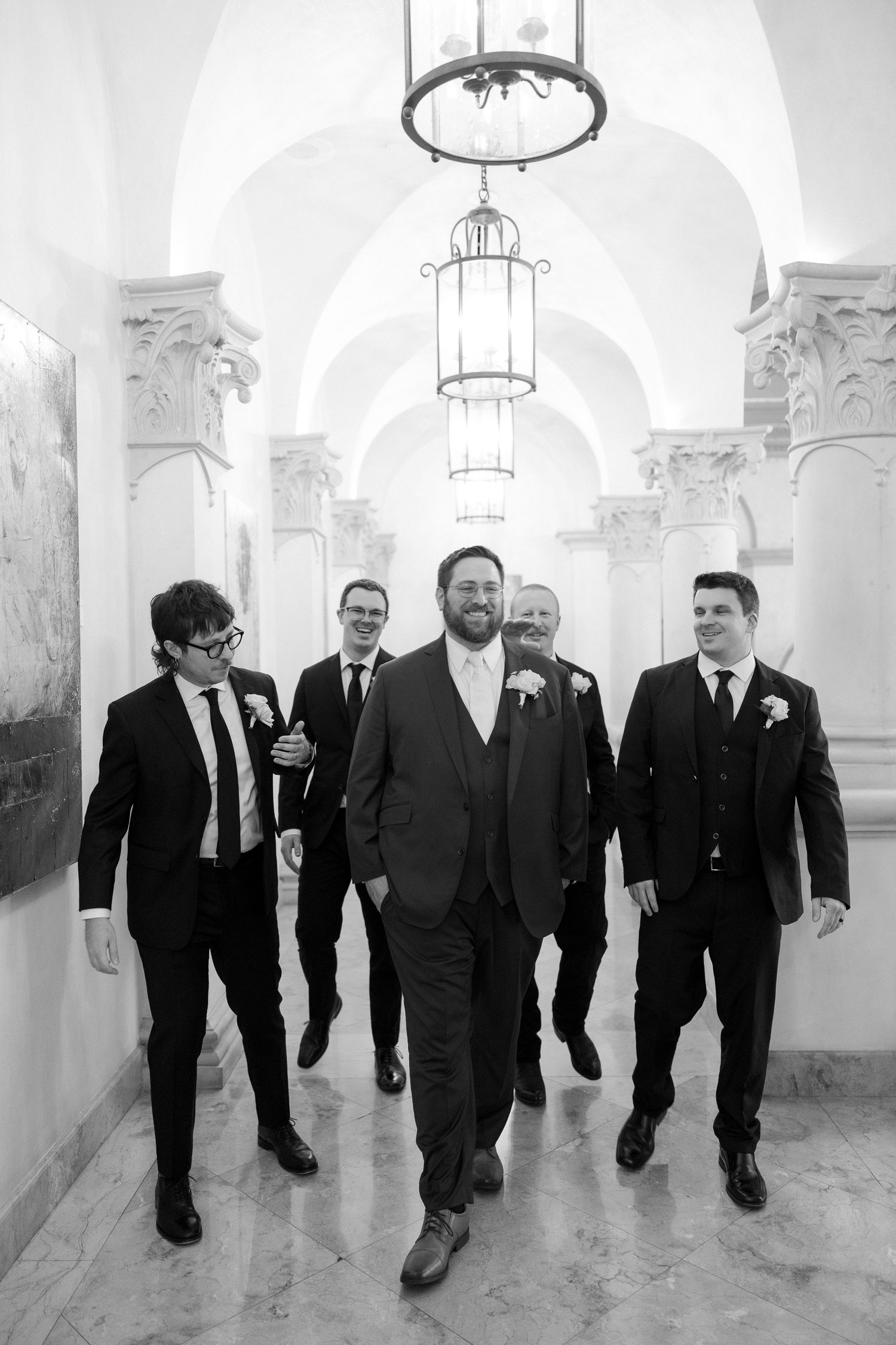Groom leads groomsmen down elegant arched hallway in black and white wedding portrait.