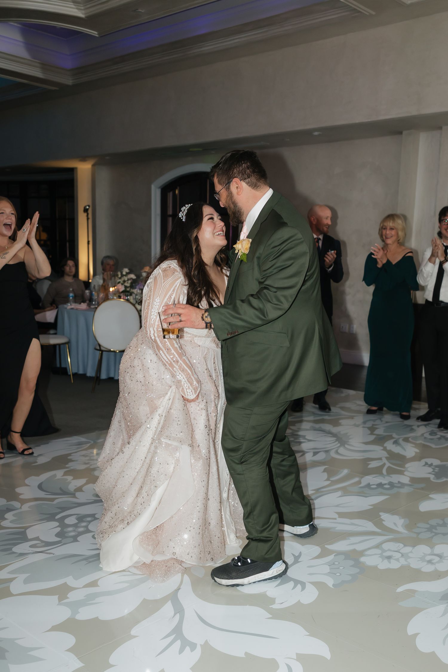 Bride and groom share first dance on floral patterned dance floor surrounded by cheering guests.