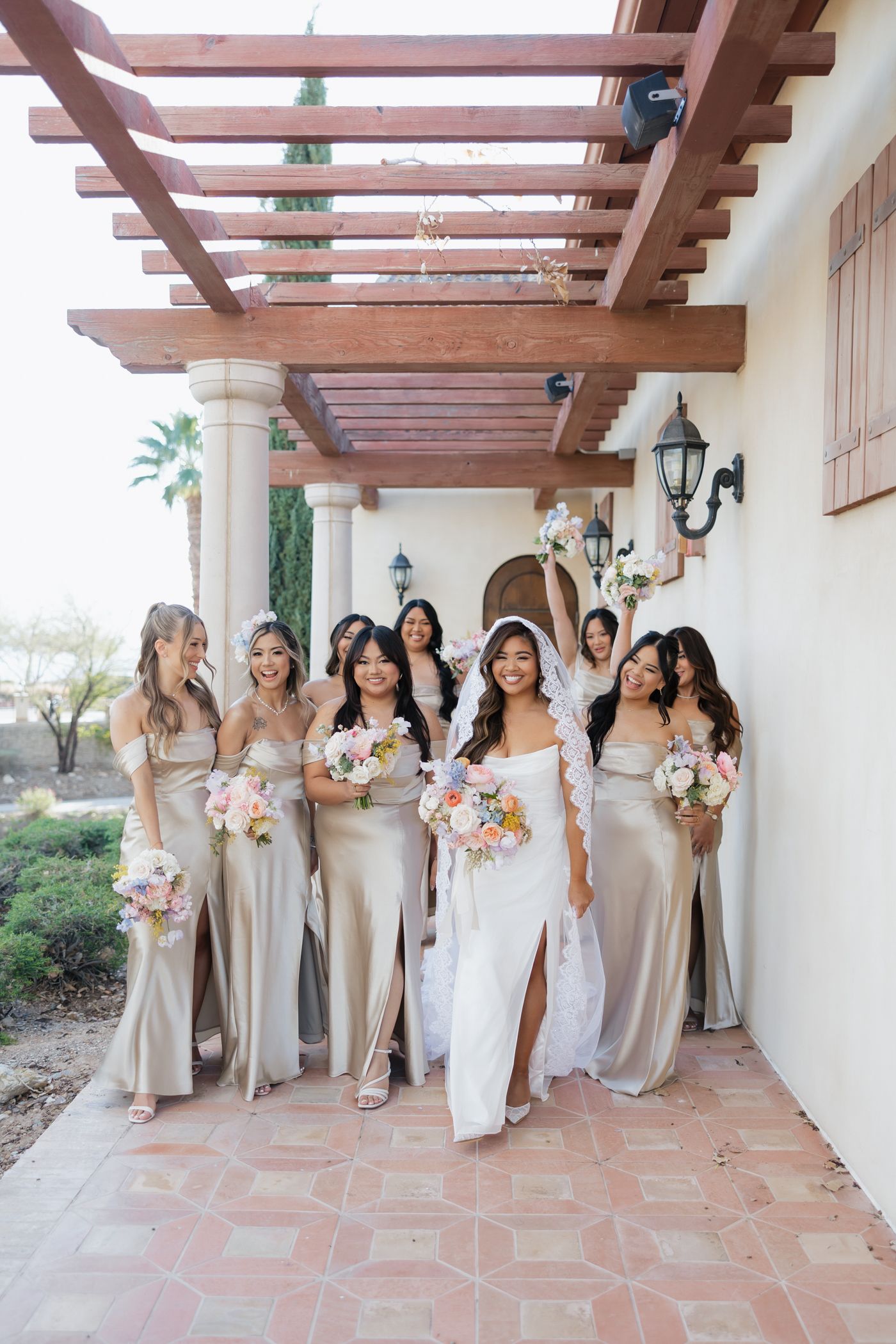 Bride in white gown walks with bridesmaids in champagne satin dresses holding floral bouquets under a wooden pergola.