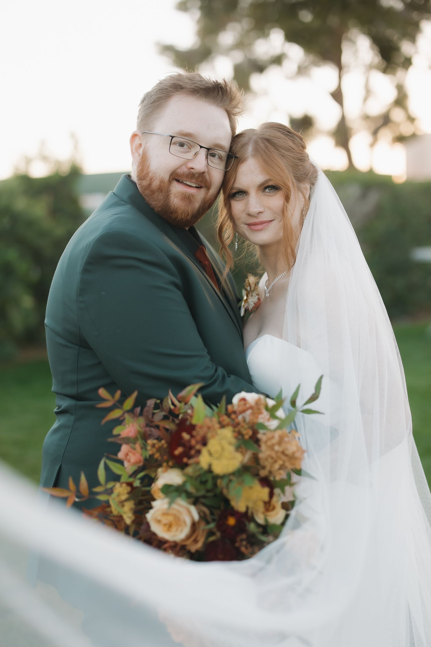 Smiling groom in green suit and bride in white veil embrace outdoors holding colorful autumn bouquet.