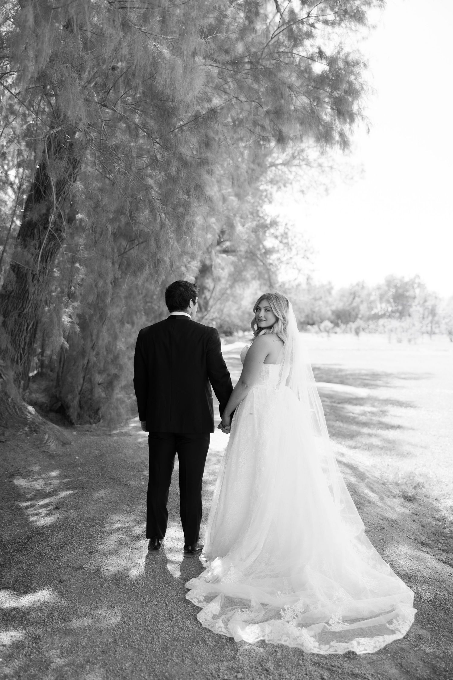 Black and white photo of a bride and groom walking hand in hand along a tree-lined path, bride glancing back.